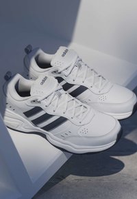 Paire de baskets blanches Adidas avec des rayures noires, lacets attachés, placées sur une surface blanche avec des ombres et un sol en béton visible.