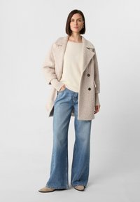 Beige strukturierter Mantel mit großen Revers und zwei schwarzen Knöpfen, kombiniert mit einem cremefarbenen Pullover und hellblauen ausgestellten Jeans. Sichtbares braunes Schuhwerk.