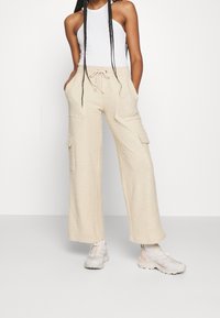 Kobieta ubrana w beżowe szerokie spodnie cargo z kieszeniami, białą koszulkę bez rękawów i pasujące beżowo-białe trampki, stoi z rękami w kieszeniach.