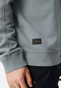 Lichtgrijze sweatshirt met geribbelde manchetten en zoom. Voorzien van een klein zwart label met gouden tekst, "MADE FOR DAILY ADVENTURES." Zachte textuur.