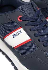 Tumesinine sneakerid, millel on võrgutextuur, valged ja punased aktsendid, logomärgiga, ümarate silmadega ja valge tallaga. Lõngad on tumesinised.