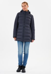 Mørkegrå dunjakke med lynlåsfront, hætte og to sidelommer. Glat overflade med polstret, quiltet design. Båret over blå jeans.