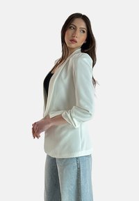 Blazer bianco con maniche lunghe, colletto a risvolto e vestibilità rilassata, abbinato a jeans in denim azzurro chiaro a vita alta. Tessuto con una texture morbida.