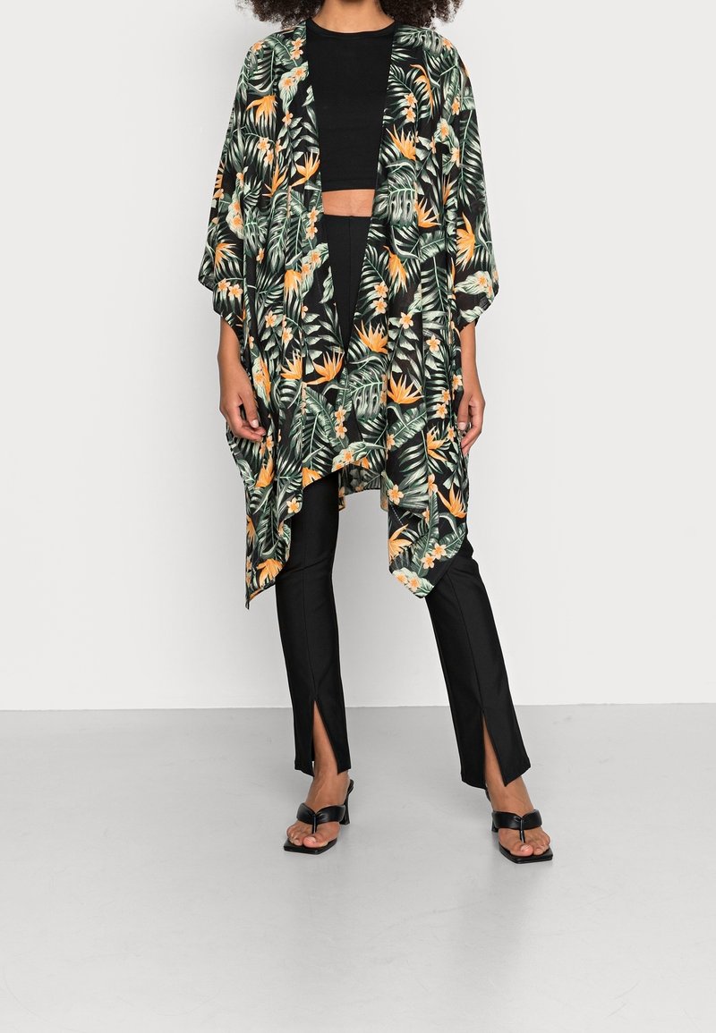 Personne portant un crop top noir, un pantalon noir à fente à l'ourlet, des sandales noires et un kimono ample à motifs floraux verts, orange et blancs.