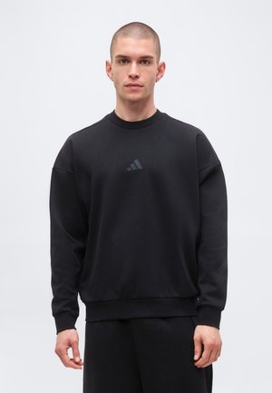 Ung mand med kort hår iført en ensfarvet sort sweatshirt med et diskret Adidas-logo, står foran en ensfarvet lysgrå baggrund.