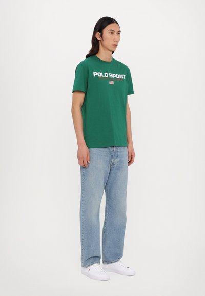 Camiseta de algodón verde con el logotipo "POLO SPORT" en blanco y acentos rojos. Combinada con unos vaqueros rectos de color azul claro y zapatillas blancas.