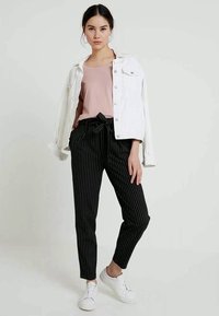 Giacca di denim bianco sopra una maglietta rosa, abbinata a pantaloni a righe bianchi e neri e sneakers bianche, con dettaglio della vita annodata.