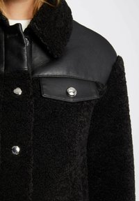 Giacca nera con una consistenza pelosa, dotata di uno yoke in pelle liscia, bottoni a pressione argentati e un design classico del colletto.