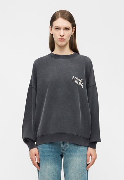 Moteris su tiesiais rudos spalvos plaukais dėvi didelio dydžio tamsiai pilką džemperį su užrašu „ANINE BING“ ant krūtinės, derinant su šviesiai mėlynais džinsais.