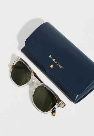 Γυαλιά ηλίου με διάφανο πλαίσιο και σκουρόχρωμους φακούς δίπλα σε θήκη Polo Ralph Lauren σε ναυτικό μπλε χρώμα με χρυσό κούμπωμα και λογότυπο, πάνω σε λευκή επιφάνεια.