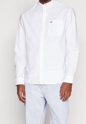 Homme portant une chemise blanche à manches longues boutonnée avec une poche poitrine et un jean bleu clair, debout devant un fond uni.