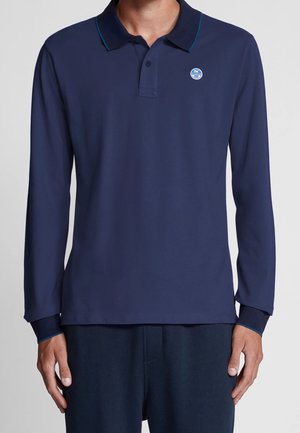 Mann trägt ein marineblaues Poloshirt mit langen Ärmeln, einem kleinen runden Logo auf der linken Brustseite und türkisfarbenem Besatz am Kragen und den Bündchen.
