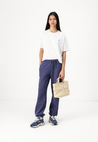 Weißes Baumwoll-T-Shirt, marineblaue Jogginghose mit elastischem Bund und blaue Sneakers. Trägt eine kleine beige Stofftasche mit Griffen.
