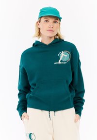 Tyrkysová mikina s predným vreckom, ktorá má krémové grafické logo. Skombinovaná s krémovými nohavicami, ktoré majú tyrkysové akcenty a detaily.