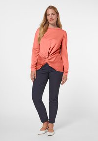 Koraalkleurig longsleeve shirt met een gedraaide voorkant, gecombineerd met donkerblauwe slim-fit broek en witte puntige ballerina's. Soepele stofstructuur.