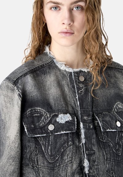 Jeune personne à la peau claire et aux cheveux bouclés mouillés, portant une veste en denim noire usée avec un col effiloché et des boutons-pression argentés.