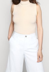 Beige geribbelde mouwloze turtleneck top, gecombineerd met witte hoge taille wijde broek met knoopsluiting en zijzakken.