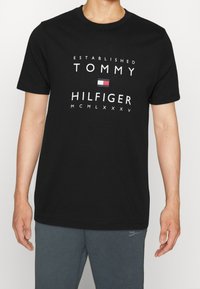 Černé bavlněné tričko s krátkým rukávem, na kterém je bílé textové označení "ESTABLISHED TOMMY HILFIGER" a malá grafika vlajky uprostřed.