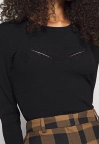 Schwarzes, geripptes Langarmshirt mit einem V-förmigen Ausschnitt nahe dem Ausschnitt, kombiniert mit einem hoch taillierten Rock in Braun und Schwarz mit Karomuster.