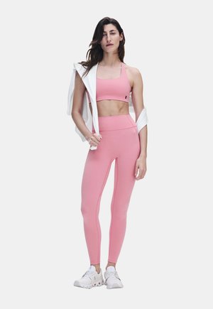 Femme debout portant un soutien-gorge de sport rose et des leggings, veste blanche drapée sur les épaules et baskets blanches sur un fond uni.