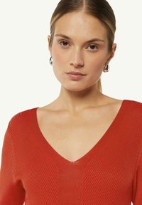 Rotes ribbel gestricktes Oberteil mit tiefem V-Ausschnitt, kurzen Ärmeln und figurbetonter Passform. Silberne Tropfenohrringe runden den Look ab.
