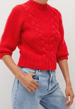 Rød strikket sweater med kabelmønstre foran og trekvartærmer, kombineret med lyseblå højtaljede jeans, hånd i lommen.