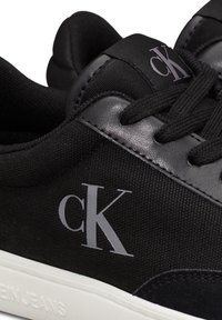 Crne tenisice od sintetičkog materijala s platnenim i nubuk detaljima. Imaju bijeli potplat i istaknuti sivi "ck" logo.