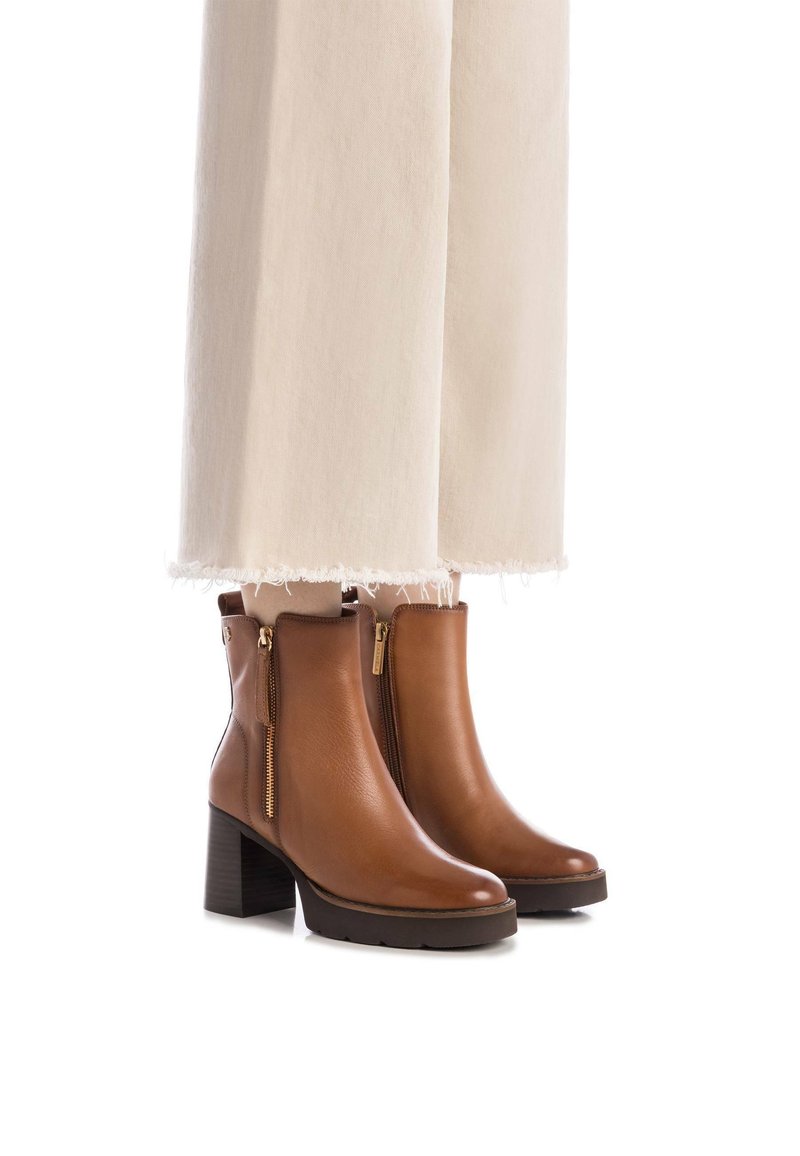 Botas de cuero marrón hasta el tobillo con cremallera lateral, tacón apilado, punta redonda y suela texturizada, combinadas con pantalones blancos con dobladillo deshilachado.