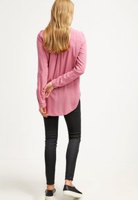 Blouse rose à manches longues avec un ourlet arrondi, des fentes sur les côtés et des détails à boutons. Associée à un jean noir ajusté et des chaussures noires à enfiler.