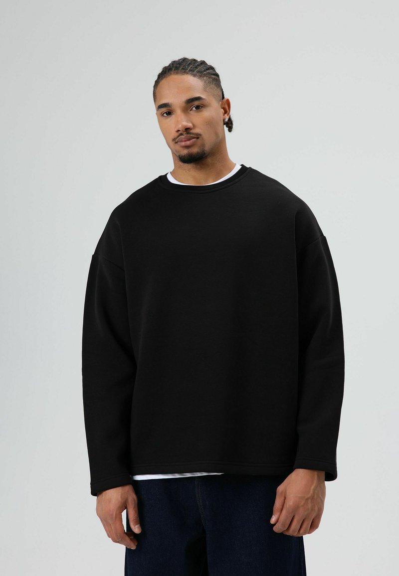 Zwart oversized sweatshirt met een ronde hals, gemaakt van gestructureerde stof. Heeft verlaagde schouders en is gedragen over een wit shirt.