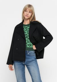 Svart peacoat med dobbelspent design, brede klaffer og knappelukking, båret over en grønn mønstret genser og blå jeans.