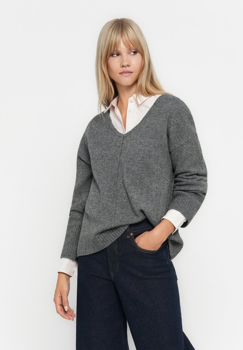 Pull en tricot gris avec un col en V et des manches longues, associé à une chemise à col rose clair. Pantalons en denim bleu foncé. Texture douce, coupe décontractée.