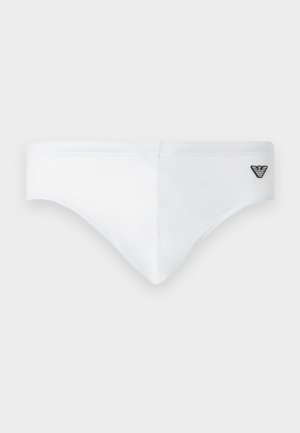 Fit man die op blote voeten staat, witte zwemslips draagt en opzij kijkt tegen een effen lichte achtergrond.