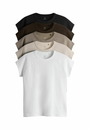 Vrouw met donker haar in een wit shirt met korte mouwen en zwarte broek staat met handen in de zakken tegen een effen witte muur.
