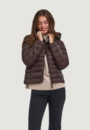 Donna che indossa una giacca imbottita marrone scuro sopra un maglione beige e jeans neri, sorridendo con gli occhi chiusi, tenendo il colletto della giacca.