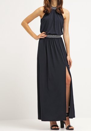Femme portant une robe maxi sans manches bleu marine avec un bord à pois au niveau de l'encolure et de la taille, dotée d'une fente haute sur la cuisse et de sandales à talons noires.