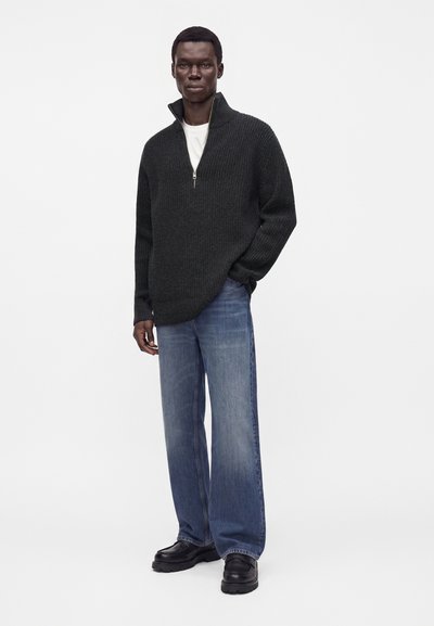 Homme debout portant un pull gris foncé à demi-fermeture éclair, un jean bleu large, des chaussures noires et une chemise blanche en dessous, sur un fond uni.