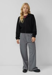 Zwarte zip-up hoodie met een getailleerde taille, gecombineerd met losse grijze broek. Glad materiaal met ribbels bij de mouwen en chunky zwarte schoenen.