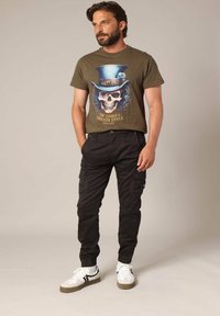 Olijfgroene t-shirt met een grafisch schedelontwerp, gecombineerd met zwarte cargobroeken en witte sneakers met grijze accenten en zwarte strepen.