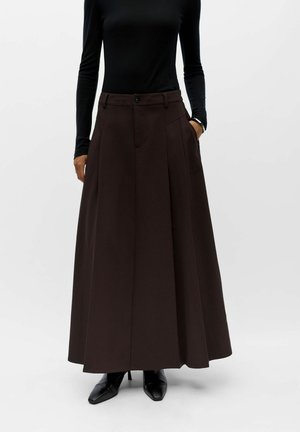 Femme portant une longue jupe plissée marron foncé, un haut noir à manches longues et des bottines à talons noires à bout pointu sur fond blanc.
