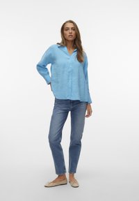 Camicia button-up azzurro chiaro in una vestibilità rilassata, con maniche lunghe e colletto, abbinata a jeans blu a gamba dritta e scarpe flat beige.