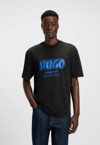 Czarny t-shirt z krótkim rękawem z wyrazistym niebieskim napisem "HUGO" oraz "CREATIVE DEPARTMENT" w białym kolorze, wykonany z miękkiego materiału.