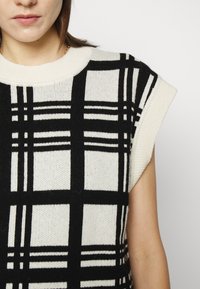 Pull en tricot noir et blanc avec un motif à carreaux, manches courtes et encolure ronde en tissu texturé.