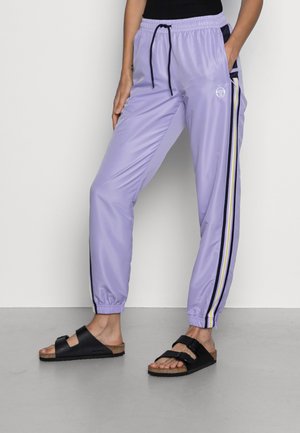 Personne portant un pantalon de survêtement violet clair avec des bandes latérales et des sandales noires, debout contre un mur et un sol gris uni.