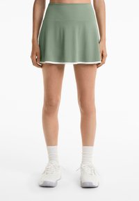 Zöld sport szoknya, bővülő szabással, puha anyagból, fehér díszítéssel az alján. Fehér tornacipővel és térdcsőig érő zoknikkal párosítva.