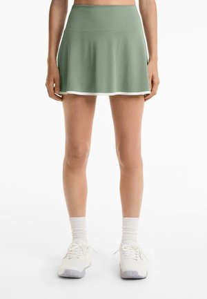 Jupe de sport verte avec un design évasé, en tissu doux, et une bordure blanche à l'ourlet. Associée à des baskets blanches et des chaussettes jusqu'au mollet.