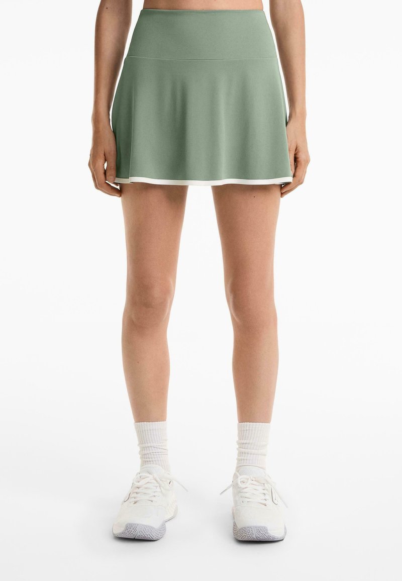 Zöld sport szoknya, bővülő szabással, puha anyagból, fehér díszítéssel az alján. Fehér tornacipővel és térdcsőig érő zoknikkal párosítva.