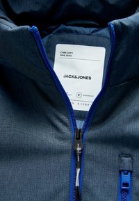 Tissu de veste bleu texturé avec une fermeture éclair bleue contrastante, étiquette avec le nom de la marque Jack & Jones, et étiquette indiquant qu'elle est coupe-vent.