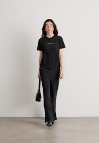 Čierne tričko s zlatým logom, skombinované s čiernymi prúžkovanými nohavicami. Modelka nesie malú čiernu kabelku a má na sebe elegantné čierne topánky.