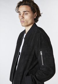 Svart mocka bomberjacka med ribbad krage, dragkedja på ärmarna och sidofickor. Mjuk textur med en figursydd siluett.